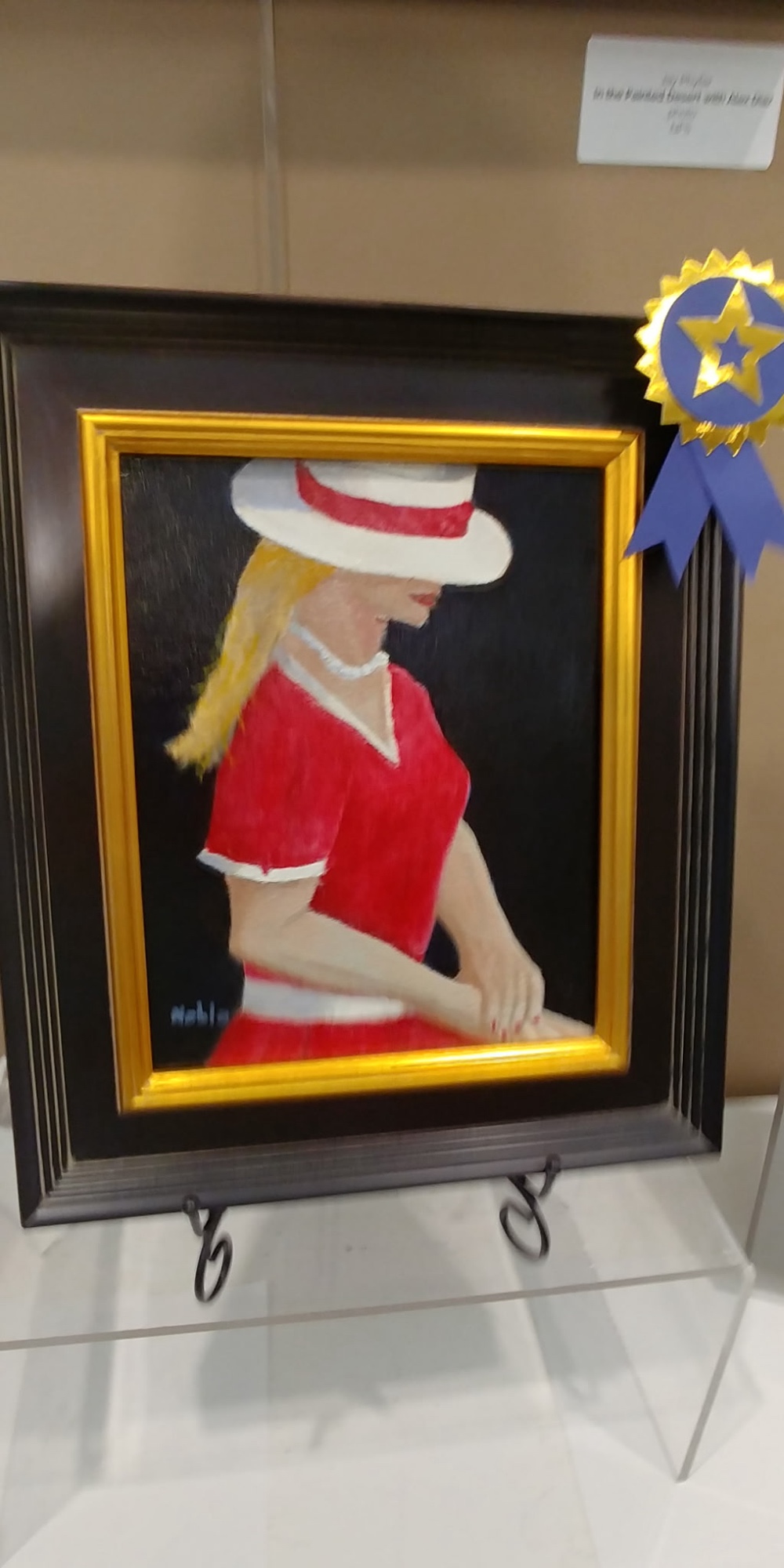 The Red Dress, a woman wearing a red dress and white hat, displayed in its frame